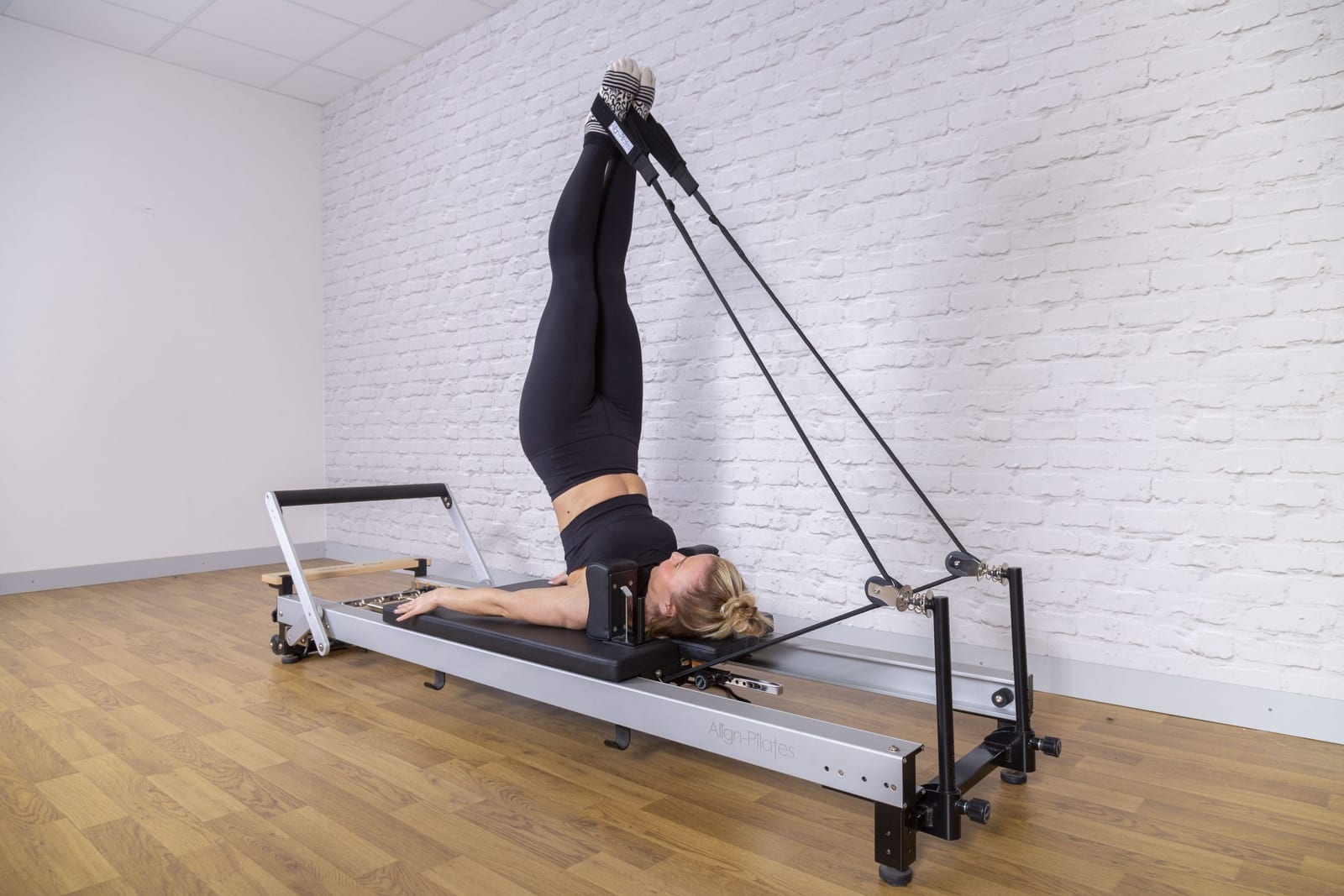 Praticante deitada no reformer com as pernas nas alças, executando um exercício de controle diante de uma parede de tijolo branco