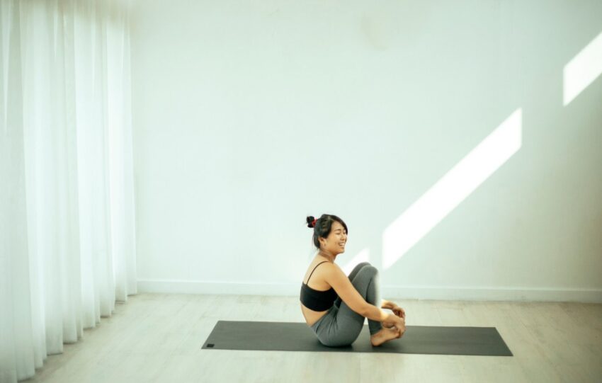 Mulher sentada sobre um tapete de exercício, com o tronco inclinado à frente e as mãos nos pés, em sala clara e vazia com luz entrando pela janela
