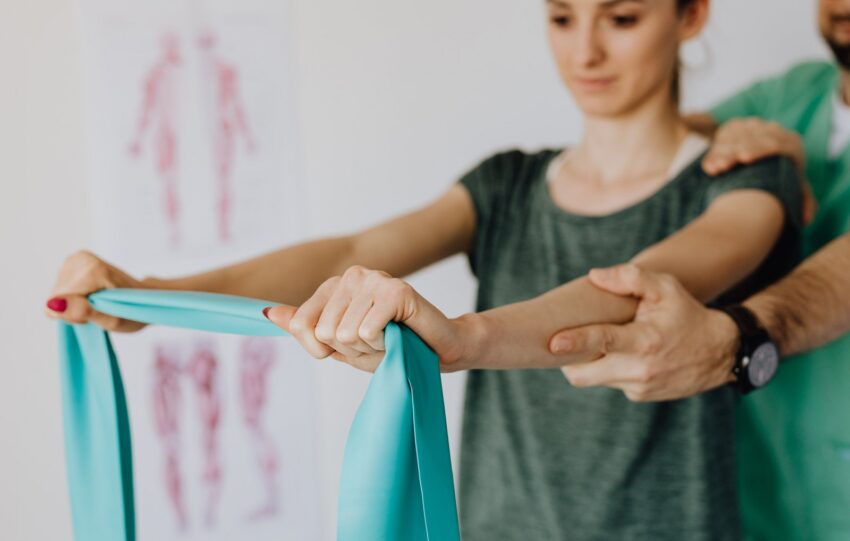 Mãos de um profissional de saúde guiando os braços de uma paciente que segura uma faixa elástica, com um cartaz de anatomia ao fundo
