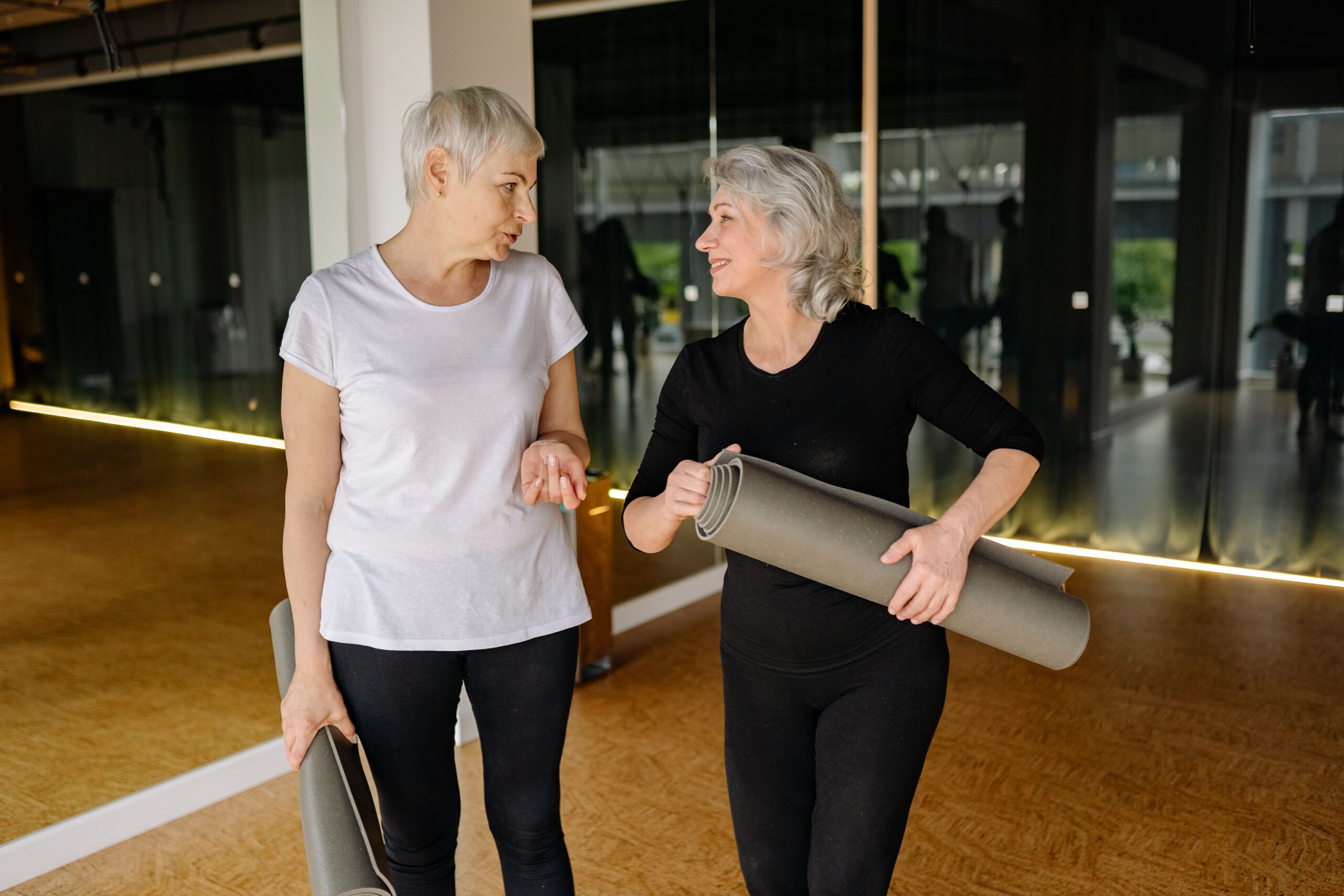 Duas mulheres de cabelo grisalho, com tapetes de exercício sob o braço, conversam de pé em uma sala de aula de ginástica, olhando uma para a outra.