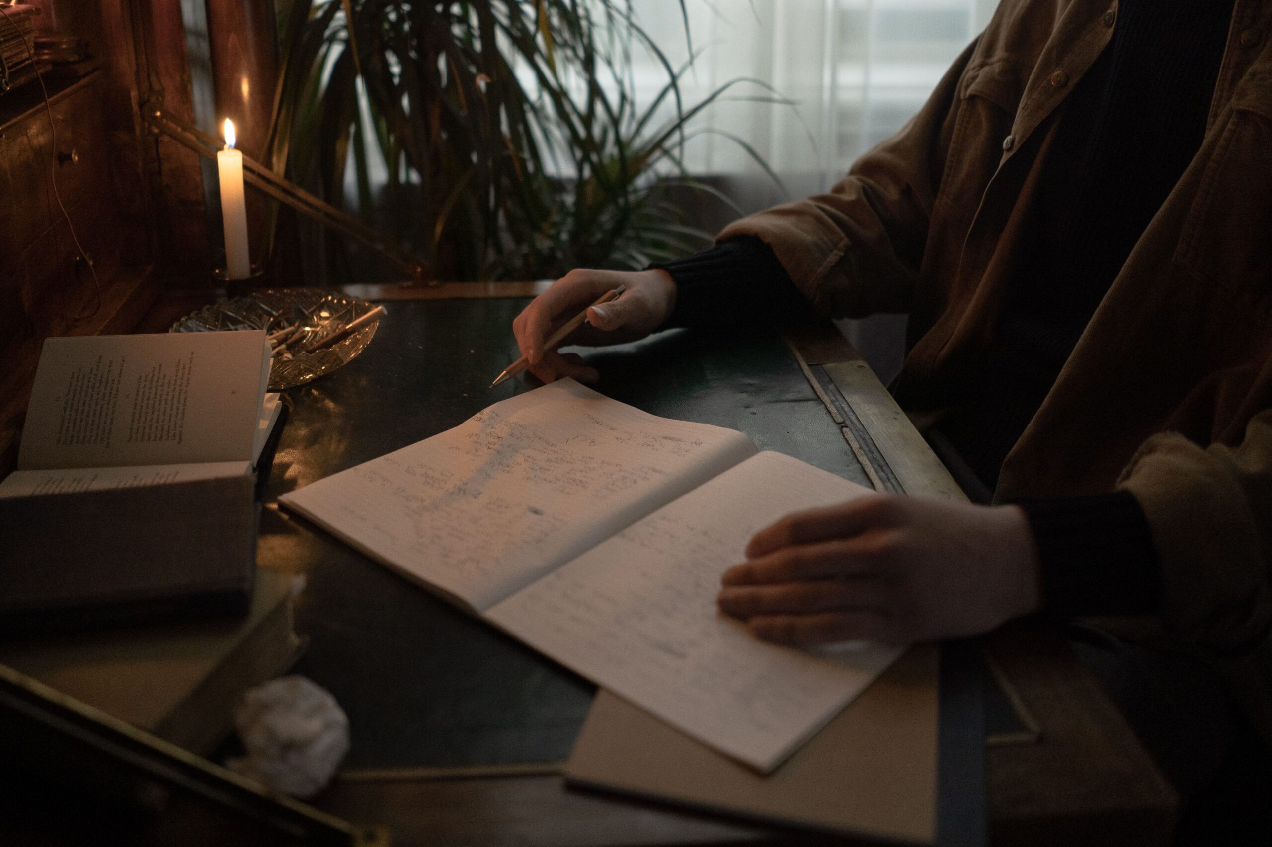 Mão segurando caneta sobre um caderno aberto com anotações manuscritas, em mesa de madeira com livros, papéis e uma vela acesa, sob luz baixa.
