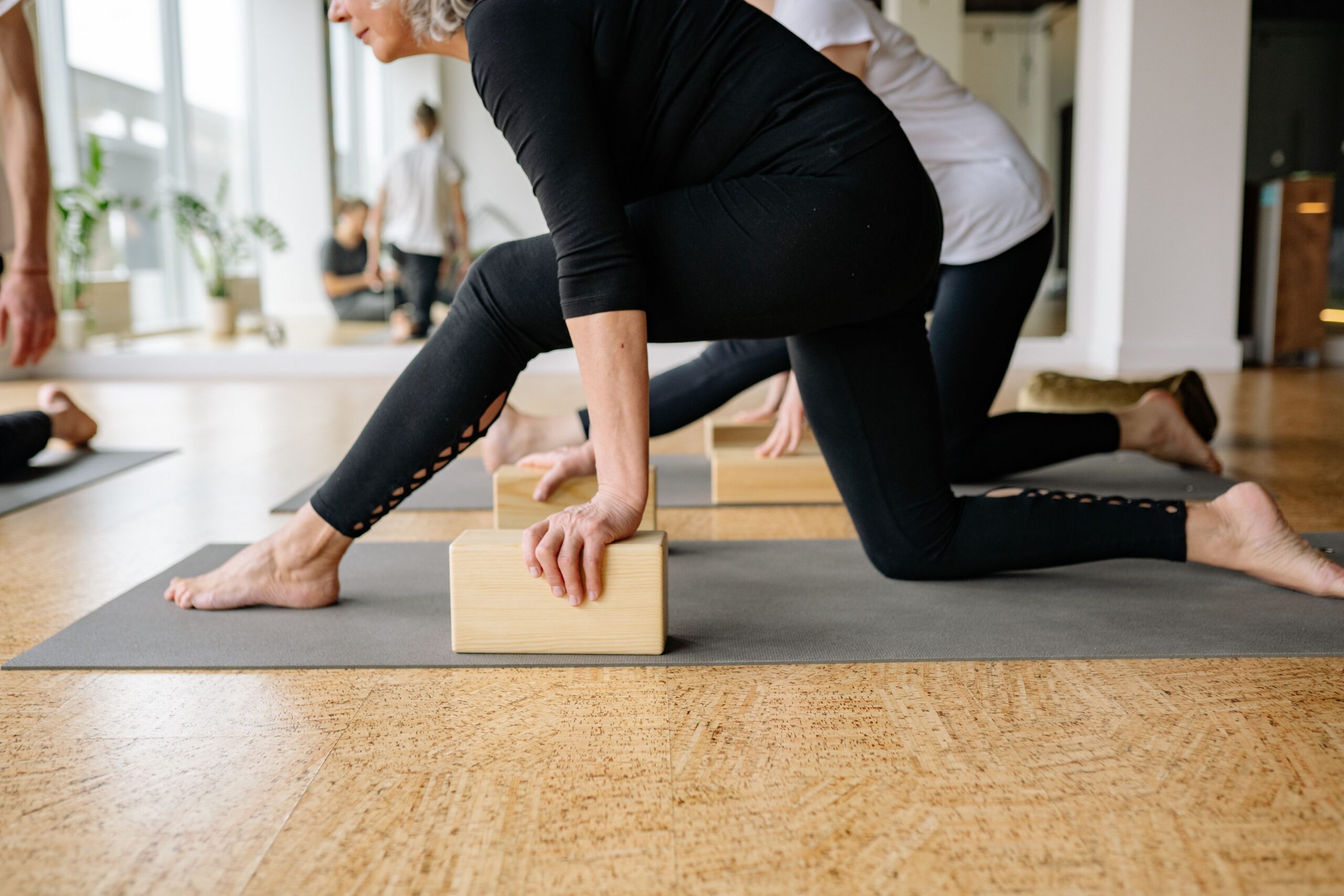 Mulheres adultas em aula de Pilates de solo sobre tapetes cinza, apoiadas em blocos de madeira, em sala com piso claro e luz de janela