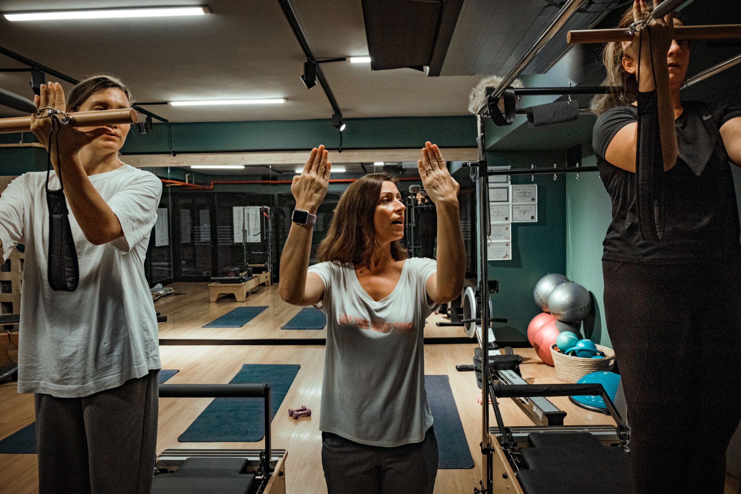 Professora de pé no meio de um estúdio de Pilates, boca aberta falando, as duas mãos erguidas com as palmas voltadas para a frente desenhando no ar a posição que ela descreve, praticantes de cada lado dela segurando barras de aparelho