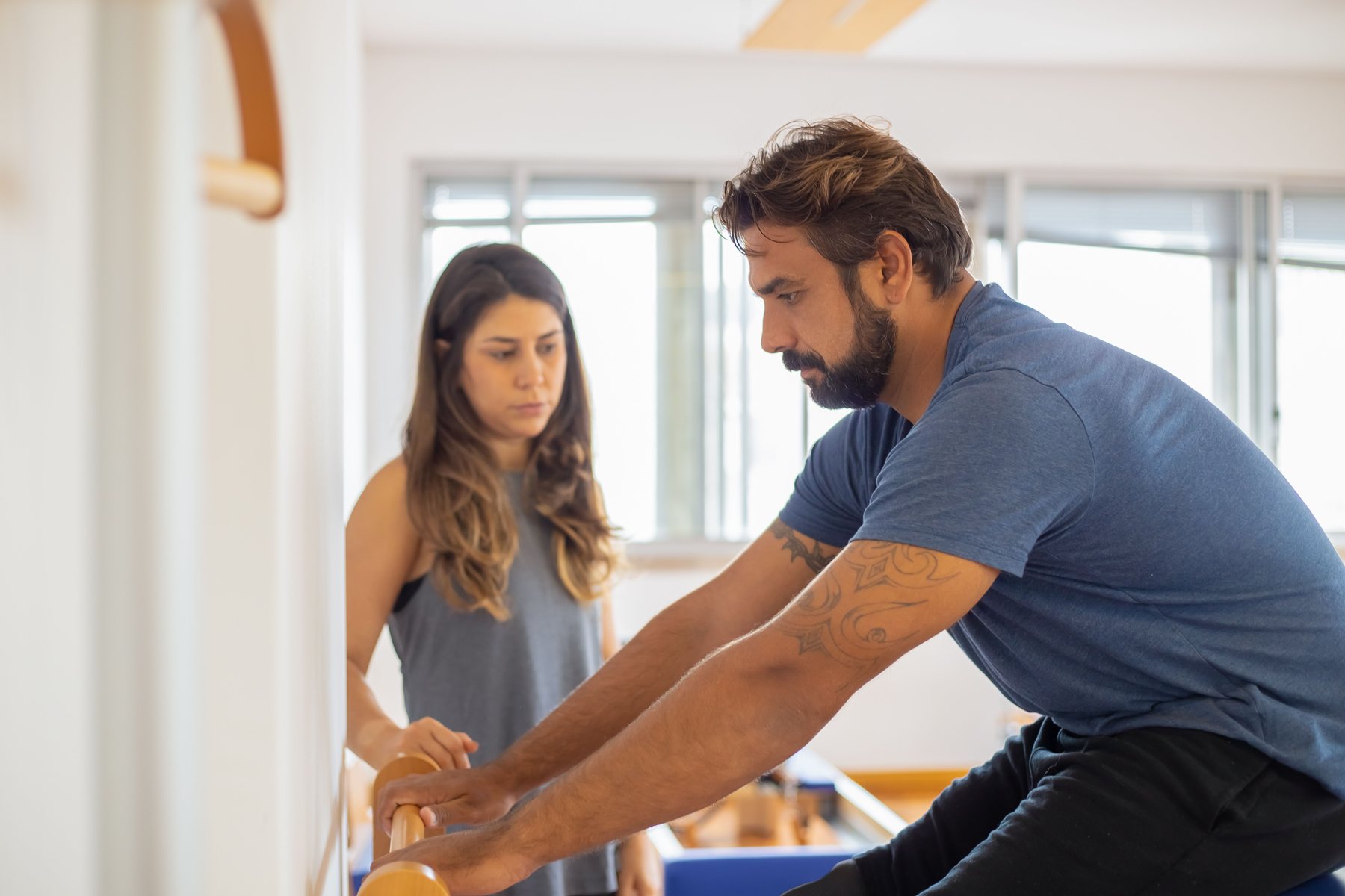 Homem de meia-idade, barba escura com fios brancos e camiseta azul, apoia as mãos numa barra de madeira em sala de Pilates iluminada por janelas, com uma instrutora ao lado observando o movimento