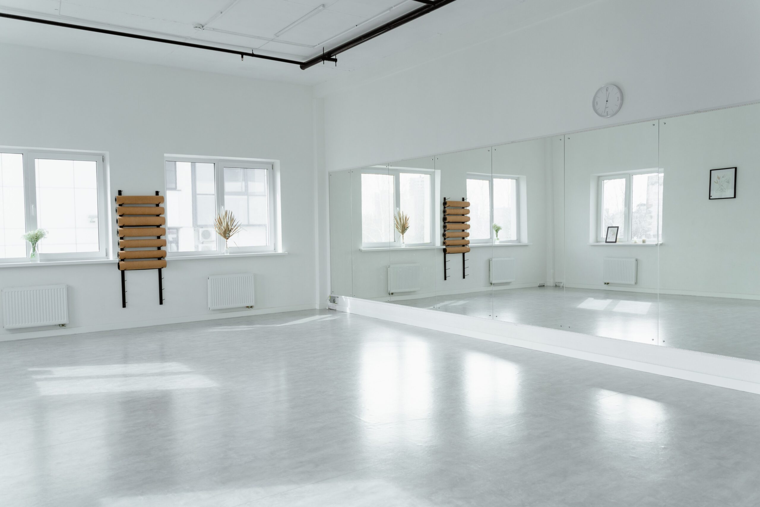 Sala ampla e vazia de estúdio, piso claro, parede branca, espelho de corpo inteiro, barra de parede de madeira, três janelas e radiadores, sem nenhuma pessoa dentro
