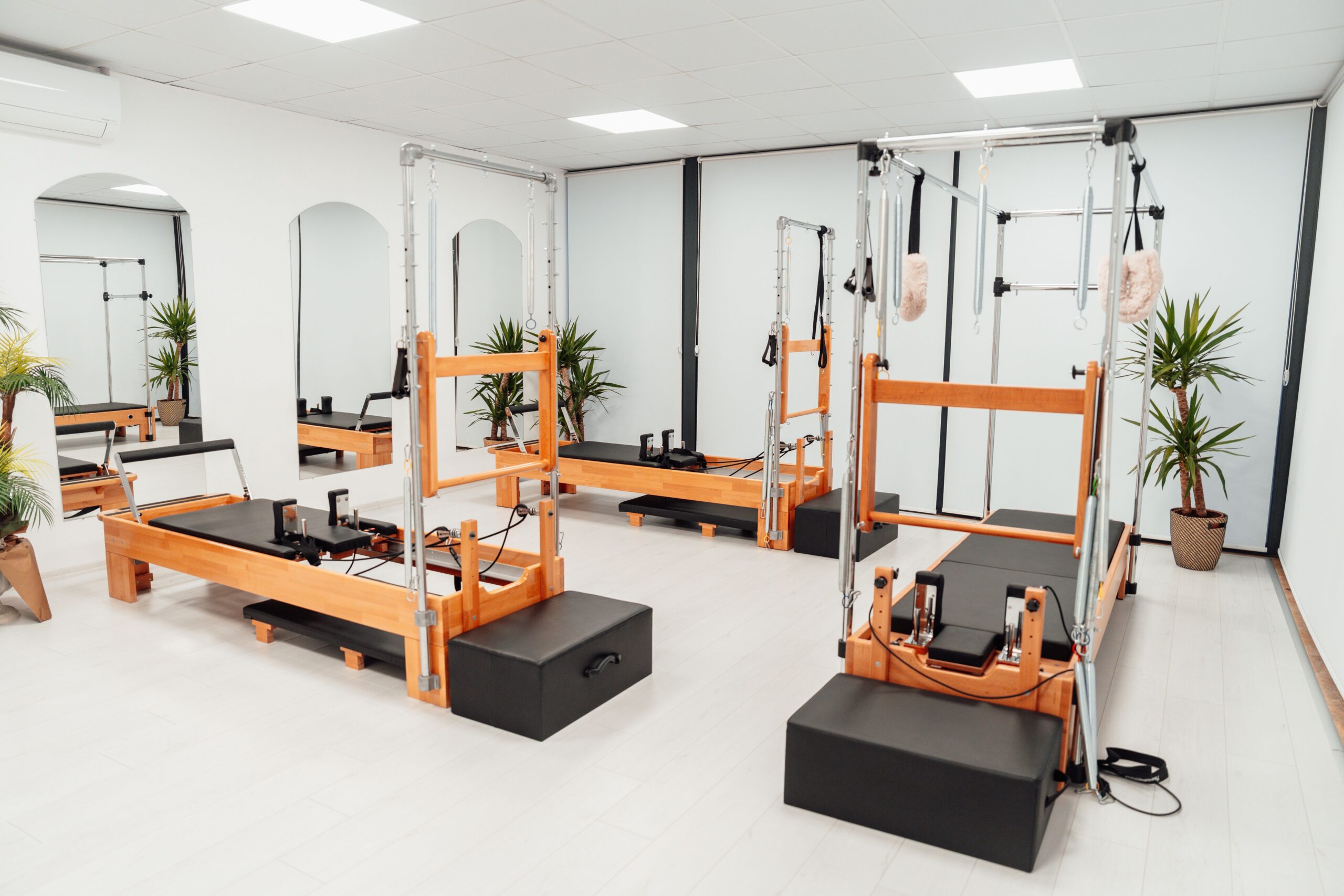 Sala de estúdio de Pilates sem ninguém dentro, três aparelhos de madeira com torre de molas parados e alinhados, piso e paredes claros, luminárias do forro acesas