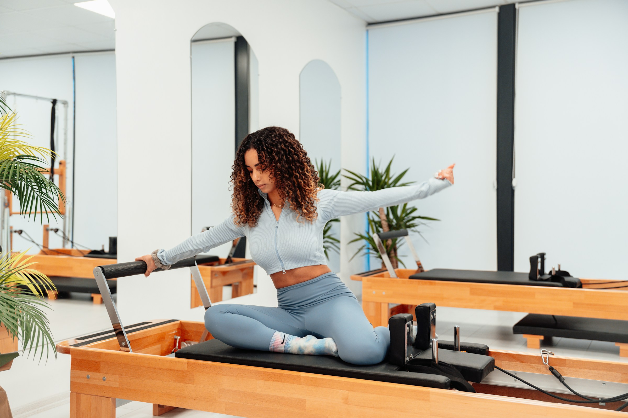 Sala de estúdio de Pilates com reformers de madeira clara idênticos e alinhados, espelhos ao fundo, e uma praticante sentada sobre um deles com um braço estendido para o lado