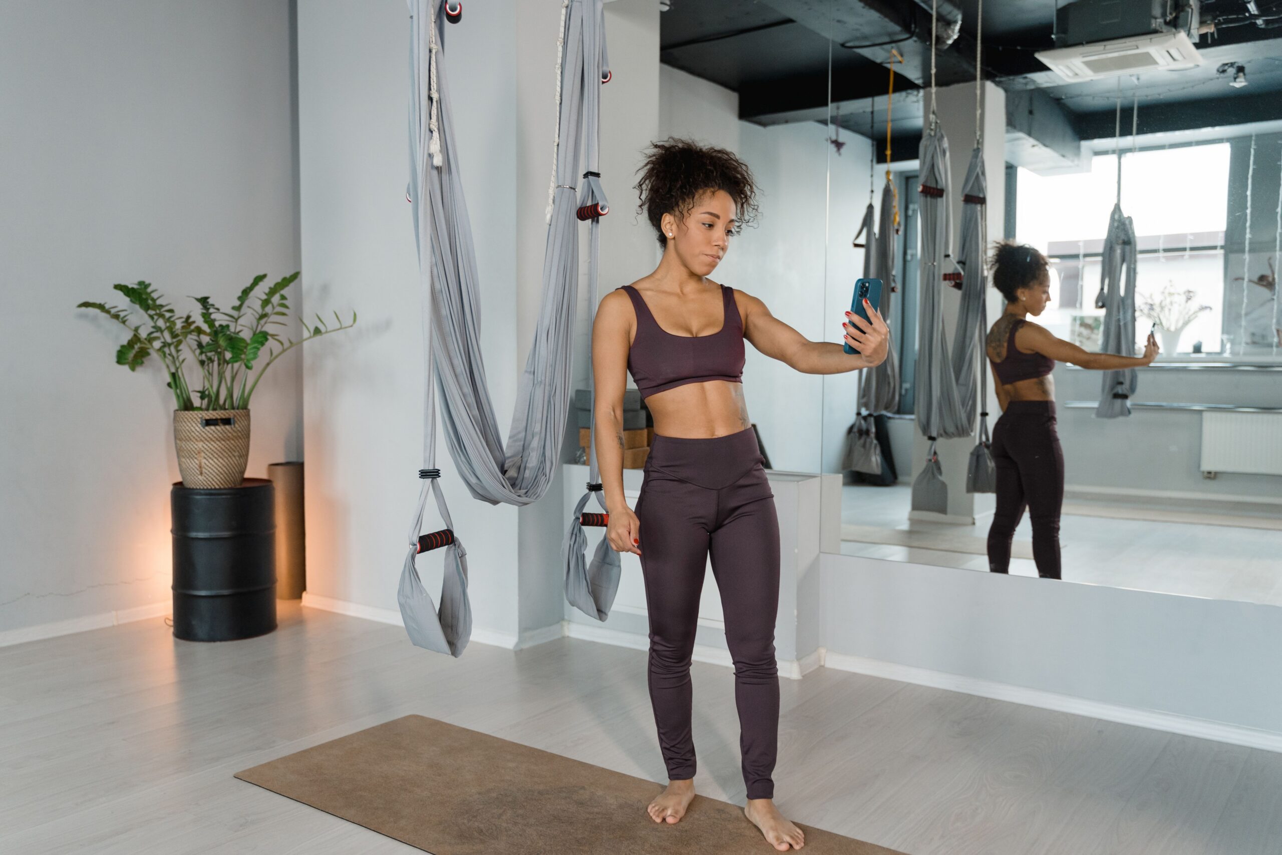Mulher de top e legging escuros, sozinha num estúdio de fitness com redes de yoga aérea suspensas, segurando um celular à frente do corpo e olhando para a tela